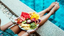 Girl relaxing and eating fruit plate by the hotel pool. Exotic summer diet. Photo of legs with healthy food by the poolside. Tropical beach lifestyle.