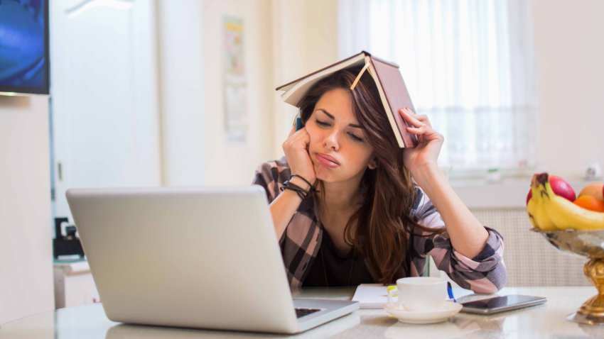 Bored female student doing homework on laptop and holding a notebook on the head.