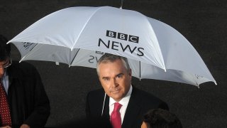 El presentador y presentador de noticias Huw Edwards sostiene un paraguas de BBC News mientras informa desde el número 10 de Downing Street el día en que Gordon Brown reemplazó a Tony Blair como Primer Ministro del Reino Unido. El corresponsal político James Landale está apenas visible a la izquierda del cuadro. 27/06/2007. Diez transmisiones exteriores de OB OBS transmiten lluvia de paraguas Número de Londres.